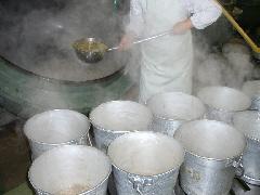 白い作業着を着たスタッフが、大きな釜を傾けてクラス別の食缶に大きなおたまで分け入れている写真