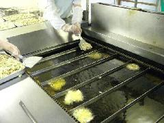 白い作業着を着たスタッフが、大きなフライヤーに材料の形を整えて油の中に投入している写真