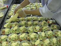 白い作業着を着たスタッフが、天板の上に並べたアルミカップに入った鮭と野菜の上にマヨネーズをかけている写真