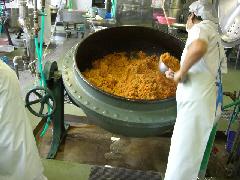 白色の作業着を着たスタッフが、大きな釜を傾けて取っ手の長い小鍋でチキンライスをすくっている写真