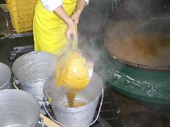 出来上がったカレールーを大きな釜からクラス別の食缶に取っ手の長い小鍋で小分けして入れている写真
