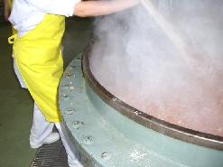 白色の作業着を着たスタッフが、湯気が出ている大きな釜に入れた材料を、大きな木べらでかき混ぜている写真