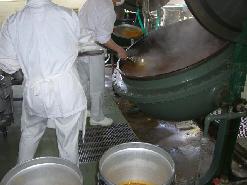 白色の作業着を着たスタッフが、大きな釜を傾けて完成したカレーを食缶に移し替えている写真