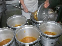 白色の作業着を着たスタッフが、クラスごとにカレーを食缶に小分けして入れている写真