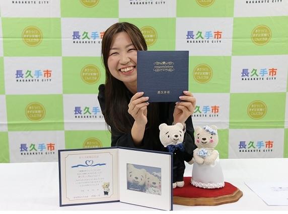 テーブルの上に長久手結婚記念証を開いた見本とタキシード姿・ウエディングドレス姿のくまの夫婦のマスコットが並べられ、その後ろで女性職員が長久手結婚記念証の表紙を見せて写っている写真