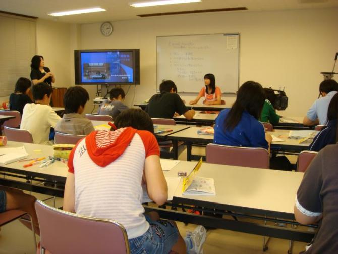 教室の前方のモニターの横に立っている1人の女性、前方のホワイトボードの前に座っている女性、訪問団OBの体験談を聞いている生徒たちの写真