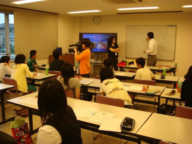 教室の前方で腕組みをしながら立っている1人の女性、ホワイトボードに書き込んでいる男性、生徒たちの様子をカメラで撮影しているケーブルテレビのスタッフの写真。
