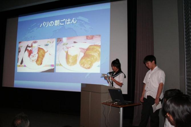 スクリーンの横に立って発表をしているAグループの女子生徒と男子生徒の写真