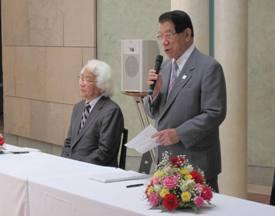 加藤梅雄町長が席から立ってマイクを持って話をしており、その横に磯見輝夫学長が椅子に座っている写真