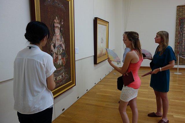 壁にかけてある大きな絵画を鑑賞しているワーテルロー市訪問団の女子学生と大学のスタッフの写真