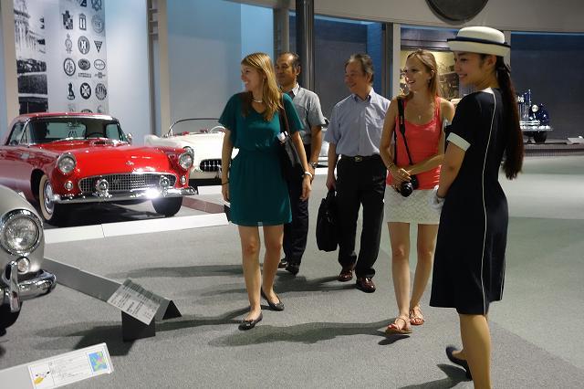 赤や白のレトロな自動車を見学しているワーテルロー市訪問団と案内の女性の写真