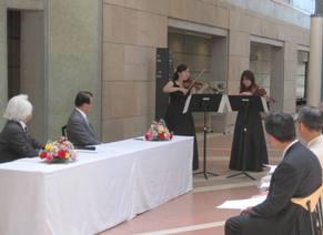 女性2名によるヴァイオリンとヴィオラによる演奏が行われており、磯見輝夫学長と加藤梅雄町長が席について、出席者も一緒に演奏を聴いている写真