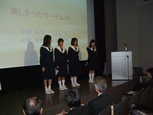 スクリーンの前に横一列に並んで立っている4人の女子生徒たちの写真