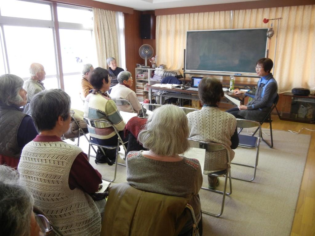 女性の講師の方が椅子に座りながら資料を手にお話をしている。椅子に座りながら参加者たちは話を聞いている写真