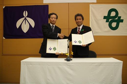 高島忠義学長と市長がサインをした協定書を広げて持って見せ、握手をして写っている写真