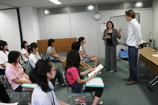 教室の前に立っている女性教師とベルギー観光局職員の男性、着席して話を聞いている生徒たちの写真