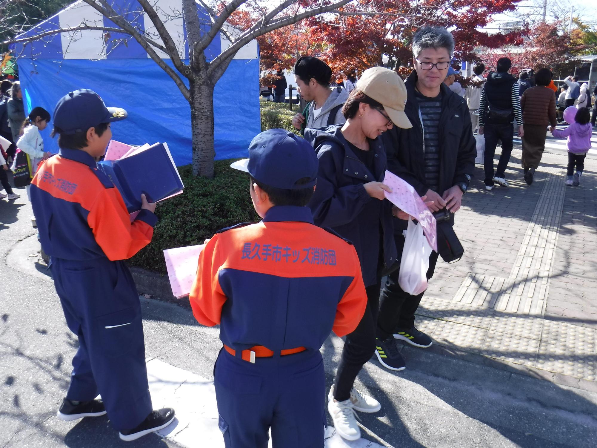 テントの裏側にある木の前で来場者にクリアファイルに入ったチラシを配る長久手市キッズ消防団の男の子たちの写真