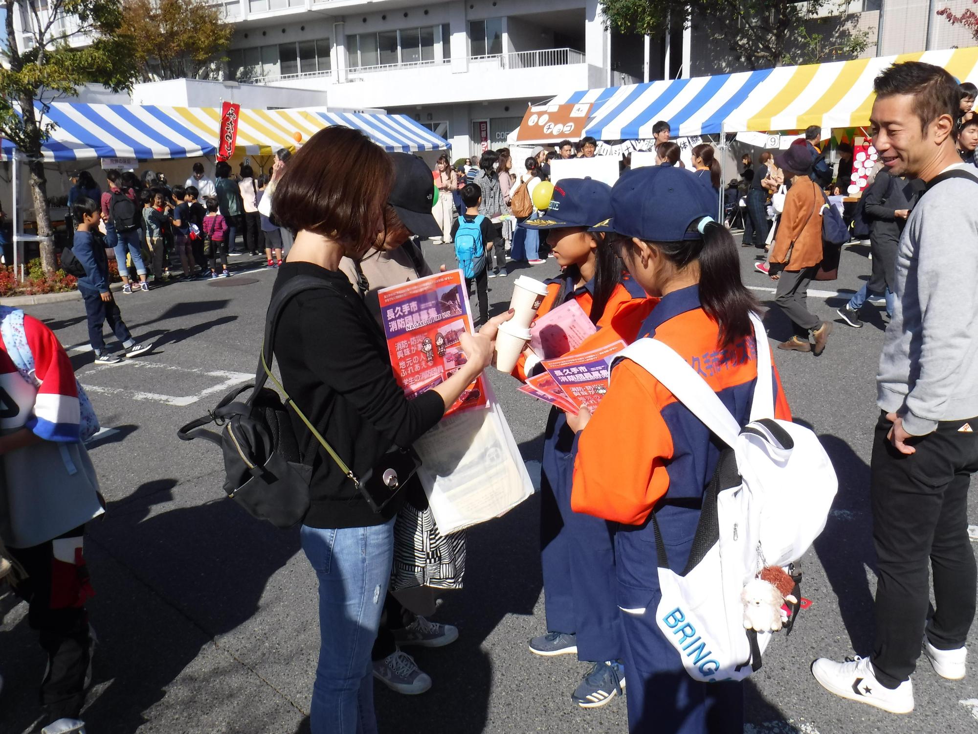 奥にテントの出店が見える市民まつりの会場で飲み物を手にしている女性たちに長久手市キッズ消防団の女の子たちがクリアファイルに入ったチラシを配る様子を見ている男性の写真