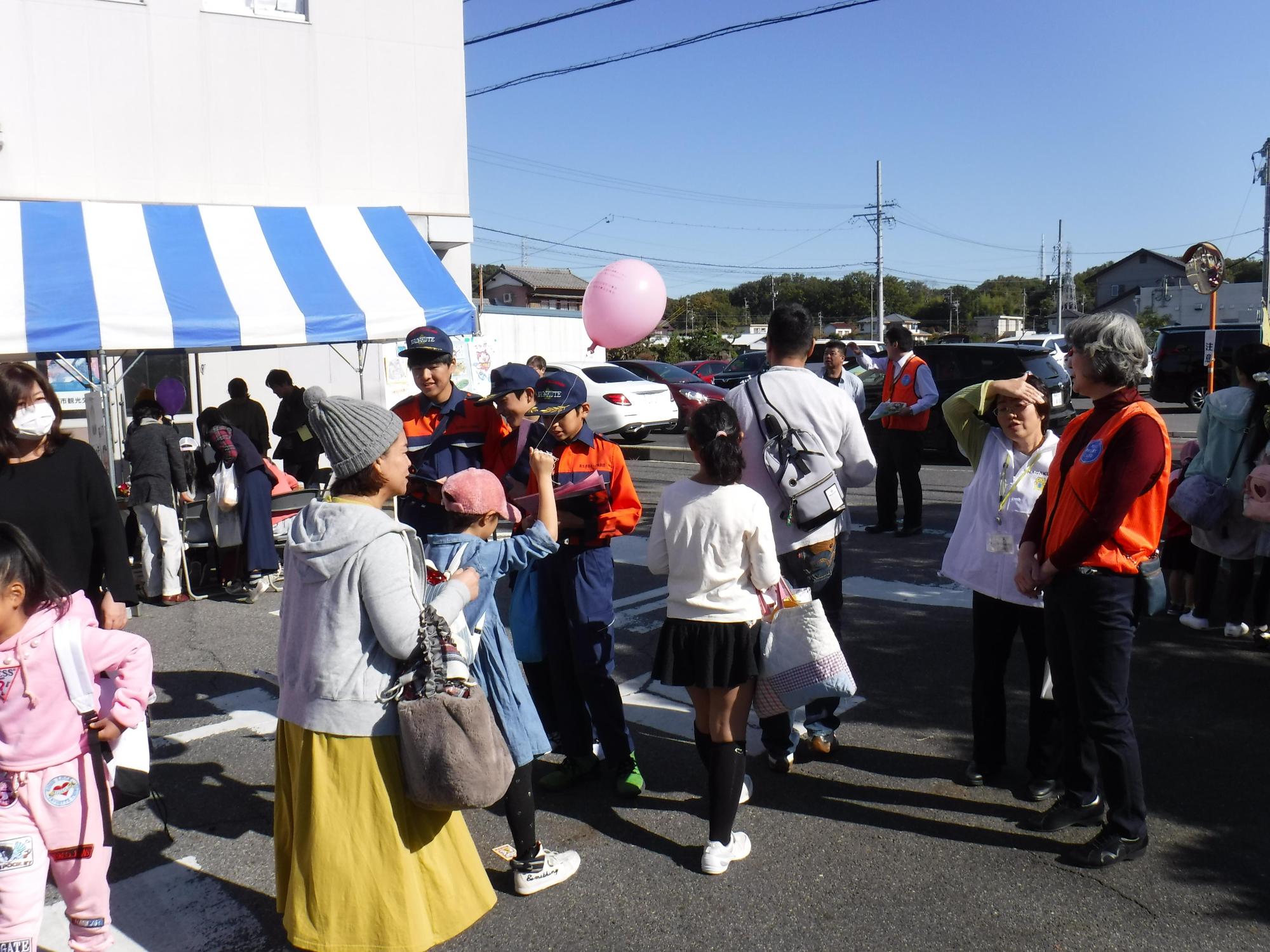 風船を持った子供や親子連れがテントの前を通っている市民まつりの会場でクリアファイルに入ったチラシを配る長久手市キッズ消防団の男の子たちの様子を見守る白やオレンジ色のベストを着たスタッフの写真