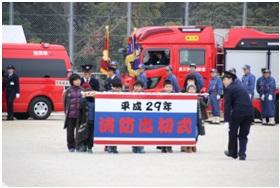消防車両が駐車されている学校のグラウンドで「平成29年消防出初式」の横断幕を持った子供たちを先頭に分列行進が行われている写真