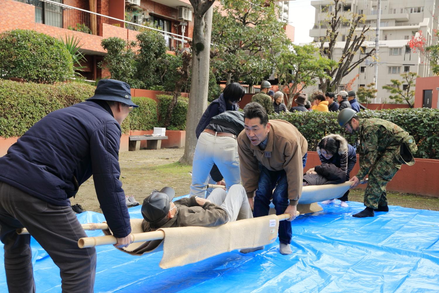 マンションの敷地内(屋外)にブルーシートが敷かれている上で竹竿と毛布で作成された担架を使って住民たちが訓練している写真