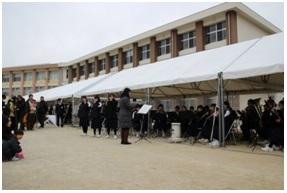 校舎の前に設置されたテントの中で先生の指揮に合わせて吹奏楽部の演奏が行われている写真