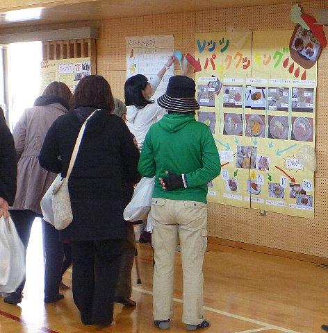 体育館の壁に、愛知淑徳大学の学生がパッククッキング紹介を大きな模造紙で作成した展示ブースを参加者が見ている写真