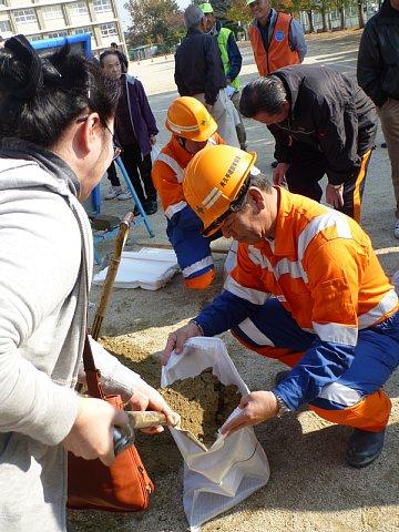 オレンジ色の作業着にヘルメットを被った男性スタッフが土のうの袋を開いているところに、女性の参加者がスコップを使って土を入れている様子の写真