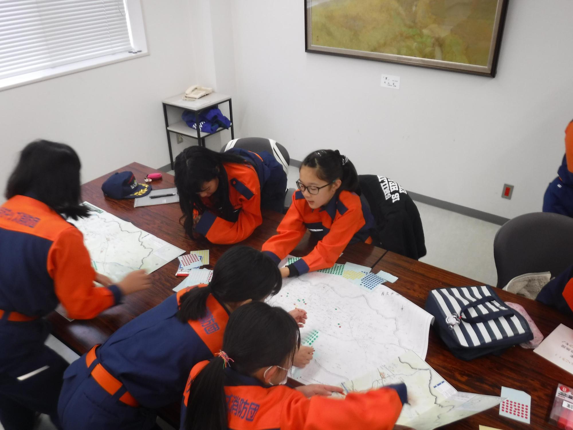 キッズ消防団員たちが地図を広げたテーブルに座り、一時避難場所、危険個所等を示すシールを貼っている写真