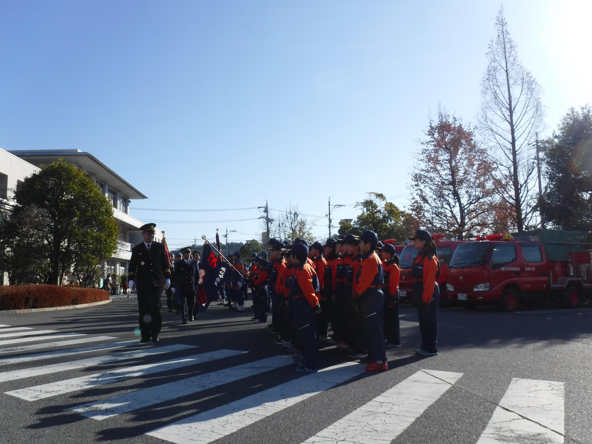 消防車両の前でキッズ消防団員たちが整列している前を団長たちが歩いている写真