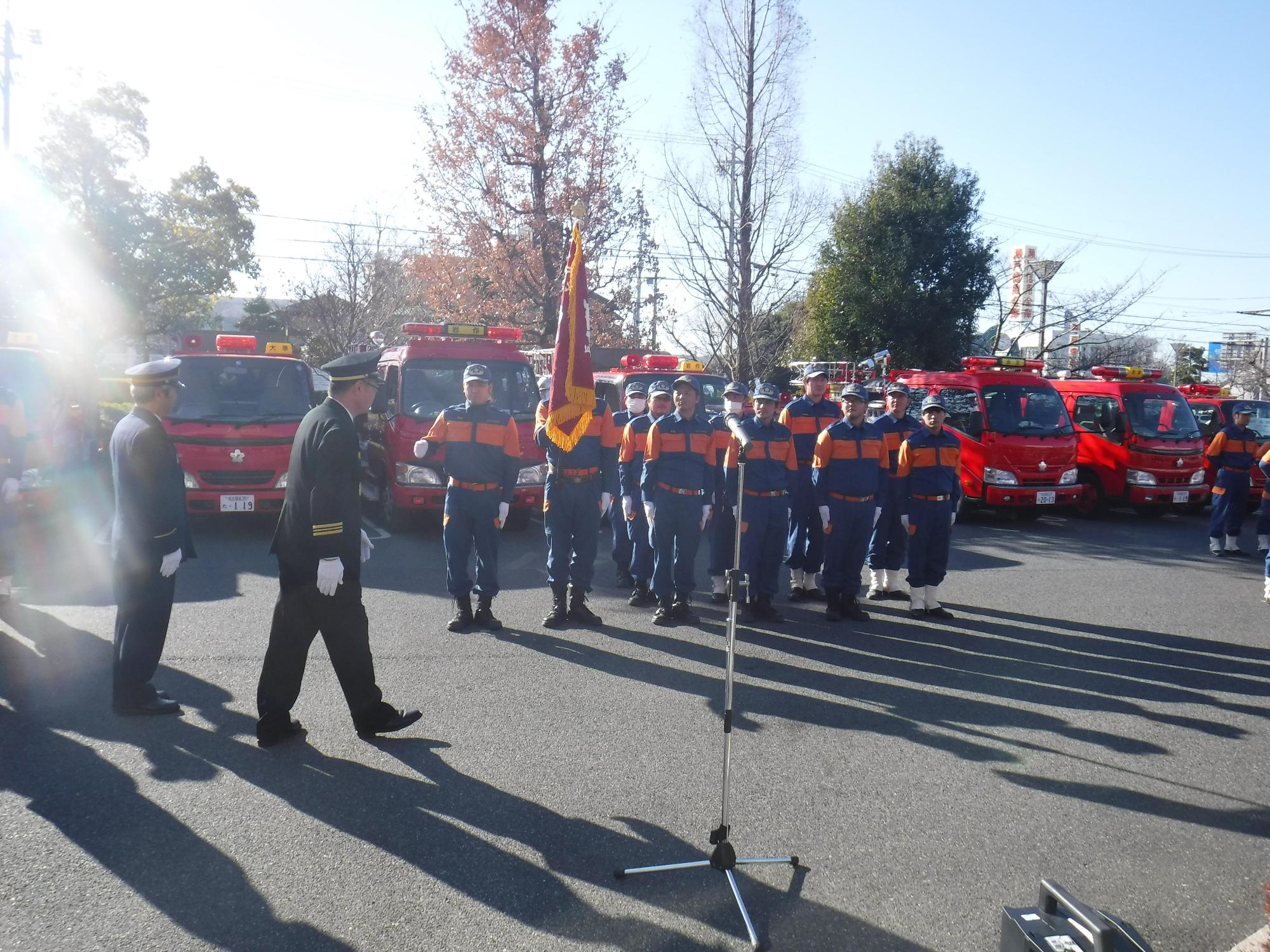 消防車両の前で消防団員たちが整列している前で挨拶しようとしている団長の写真