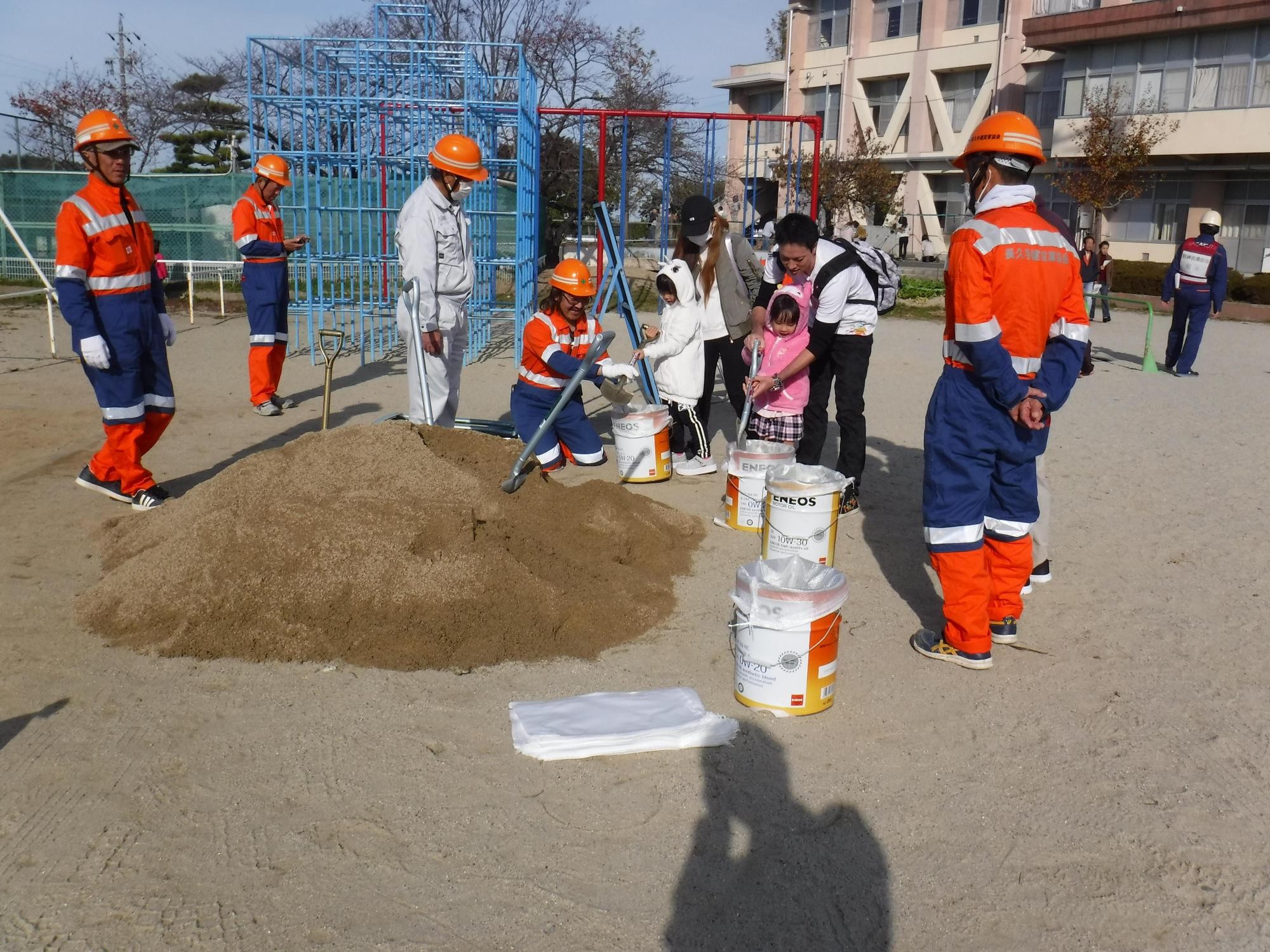 校庭のジャングルジムの前に砂が用意されており、消防団と一緒に土のう袋にスコップで砂を入れる参加者親子たちの写真