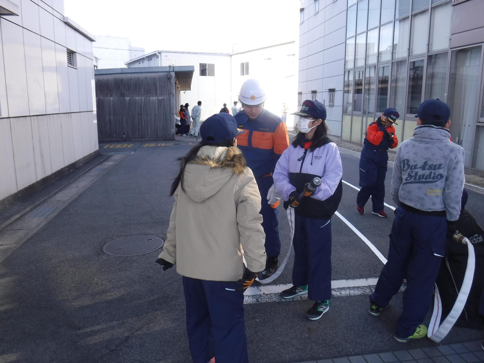 消防団員の指導のもと、建物の前でホースの延長やホース巻の訓練を行う長久手市キッズ消防団の写真
