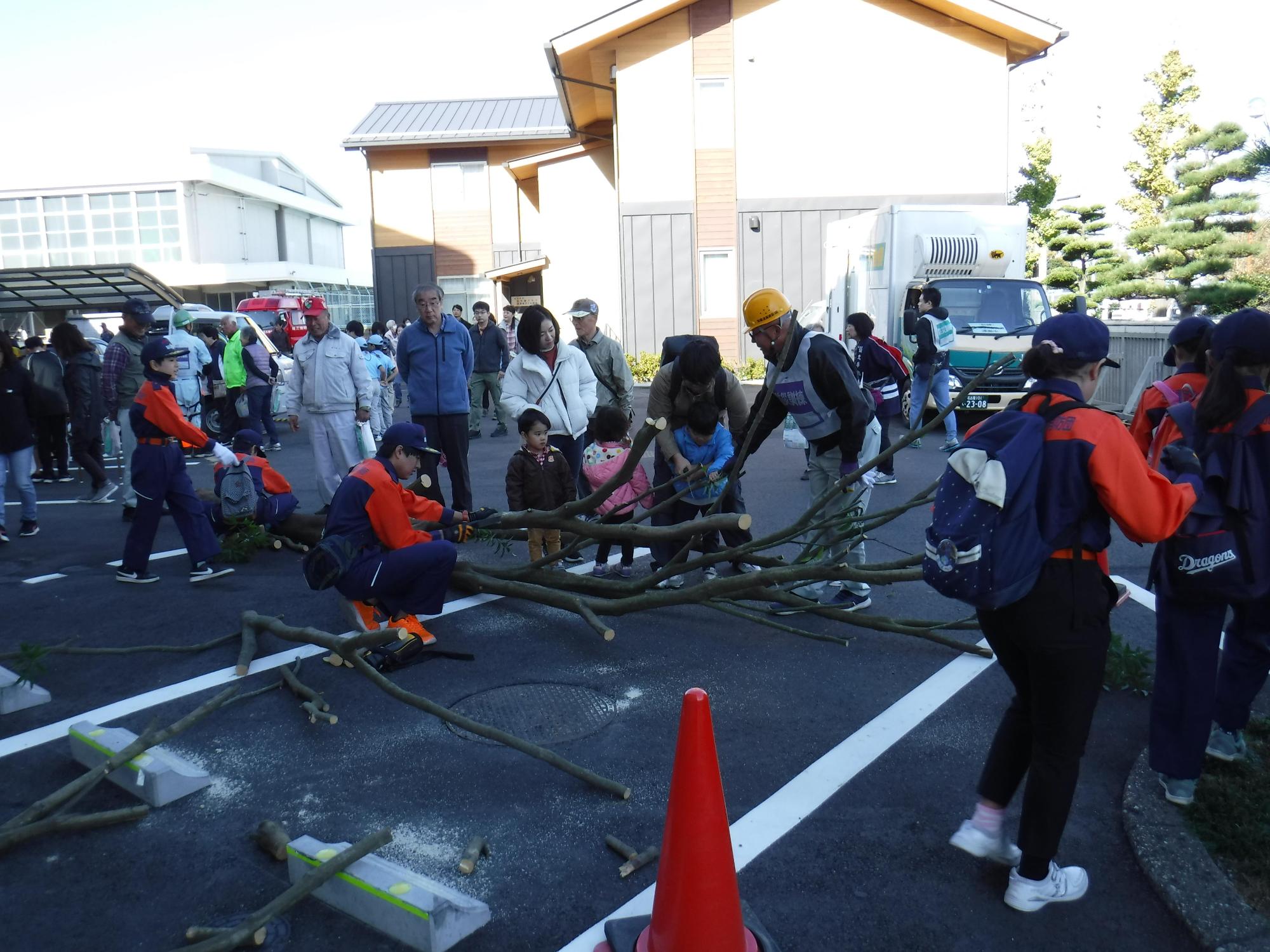 学校の駐車場に大きな木の枝が置いてあり、参加者たちが消防団員の指示に従って訓練を受けている写真