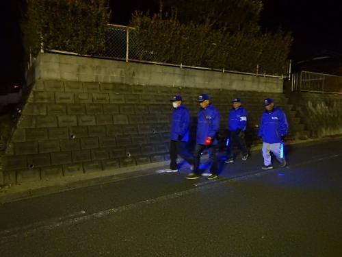 青い帽子に青色のジャンバー姿の西小校区防犯コミュニティ4名の人たちが青いLEDライトを持って夜間パトロールをしている写真