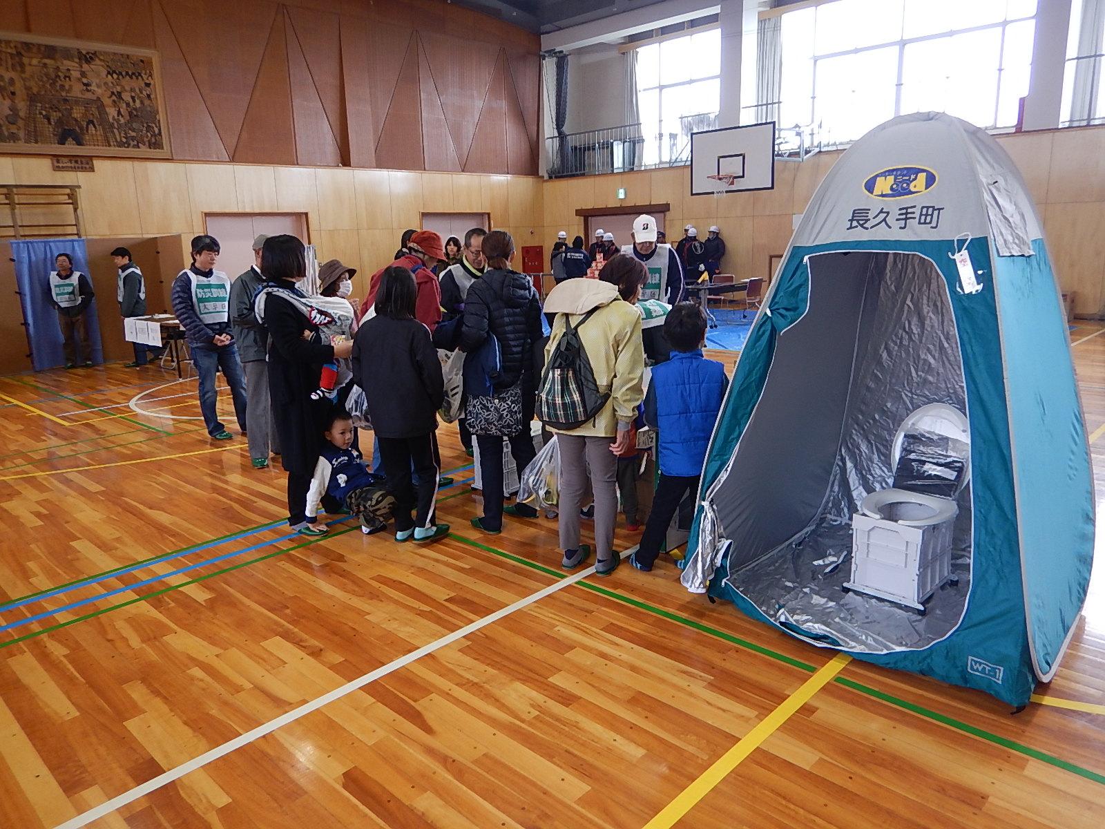 体育館内でリュックを背負った避難訓練の参加者が集まっている横にドーム型テントに設置された簡易トイレが展示されている写真