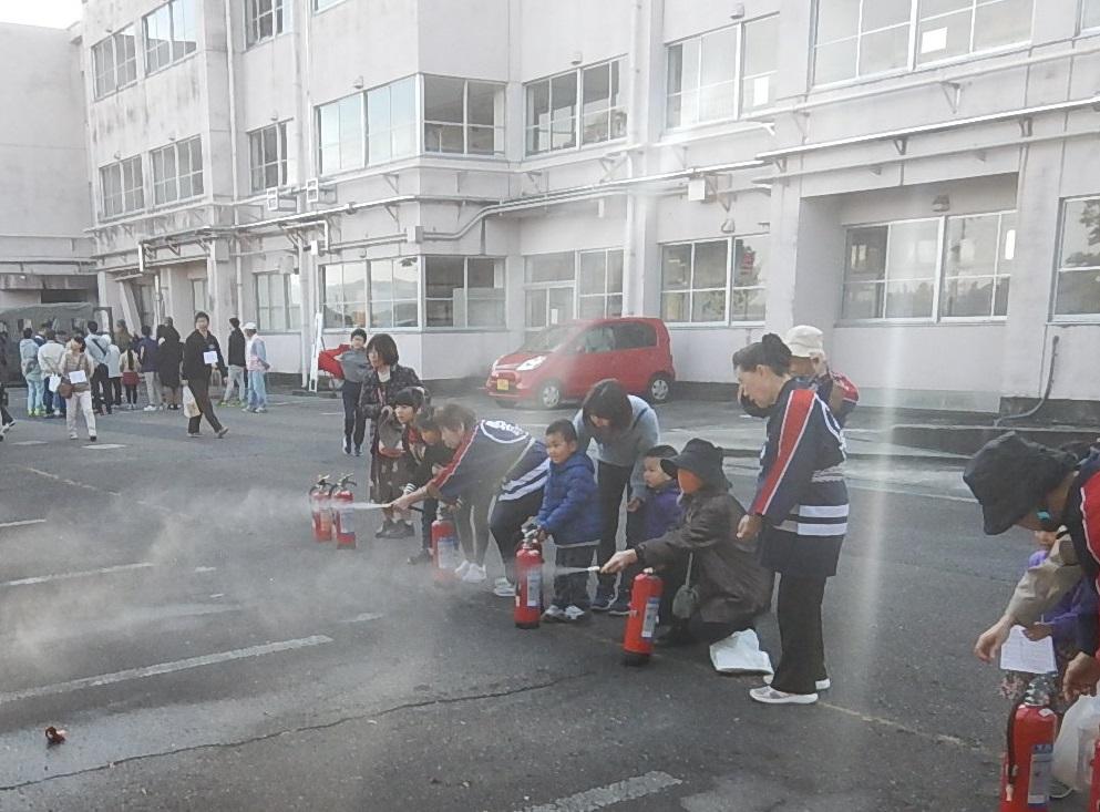 学校の駐車場で法被姿の女性消防クラブのスタッフと一緒に参加者の親子たちが消火器の放射をしている写真