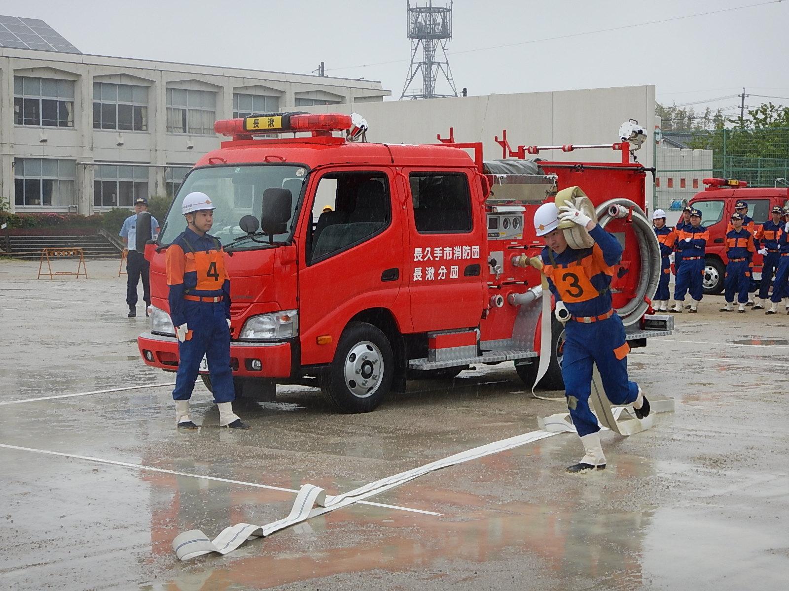消防操法大会にて消防車両の前をゼッケン3を付けた団員が放水ホースを担いでいる様子をゼッケン4を付けた団員が見ている写真