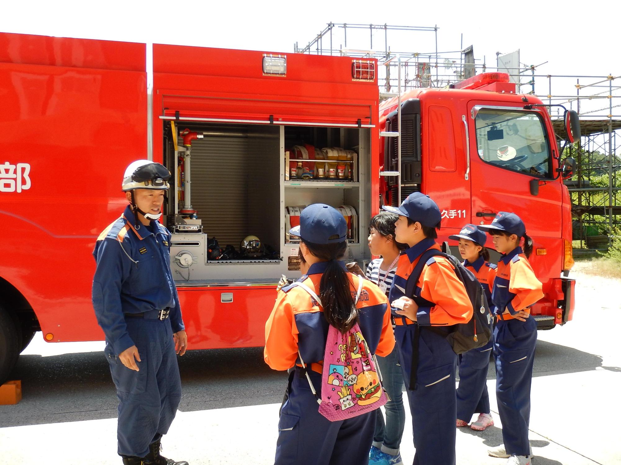 消防車両の前で消防団員からの説明を受けるキッズ団員たちの写真