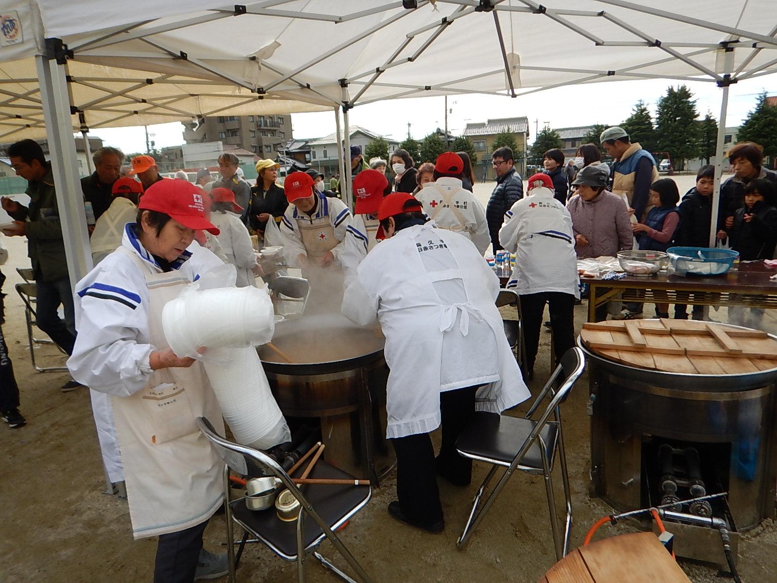 テントの下で赤い帽子を被り白いエプロンを付けた人々が大きな鍋で炊き出しを行っている写真