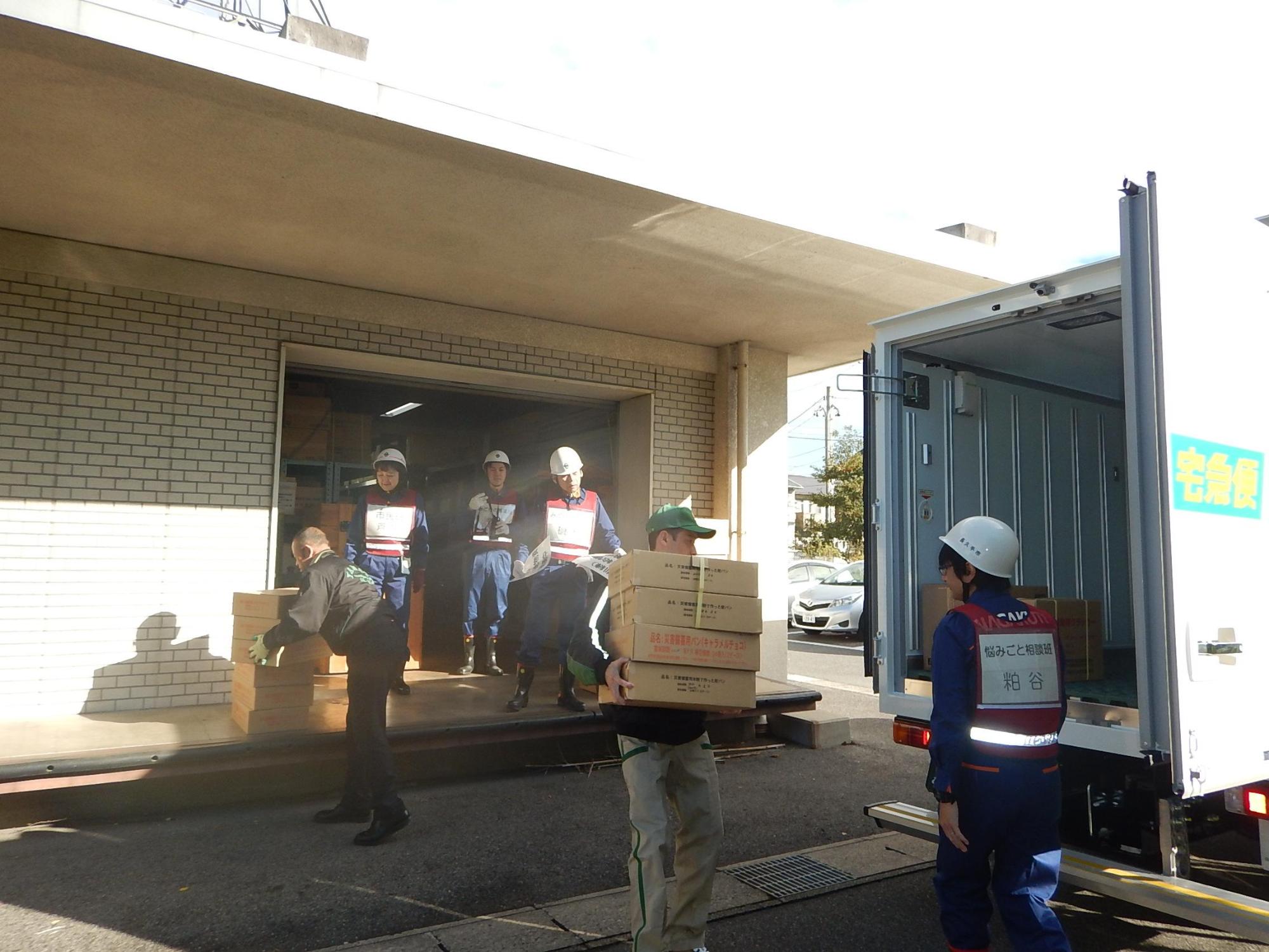 市防災倉庫前で緑の帽子を被ったヤマト運輸株式会社のスタッフが段ボールの物資をトラックに運ぼうとしている様子を青い作業服にヘルメットを被った市職員たちが見ている写真