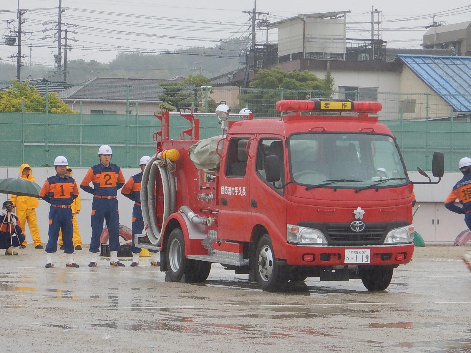 雨の降るグラウンドに消防車両が停まっており、消防車両の後ろで白いヘルメットを被りゼッケンをつけた消防団員が手を後ろに組んで立っている写真