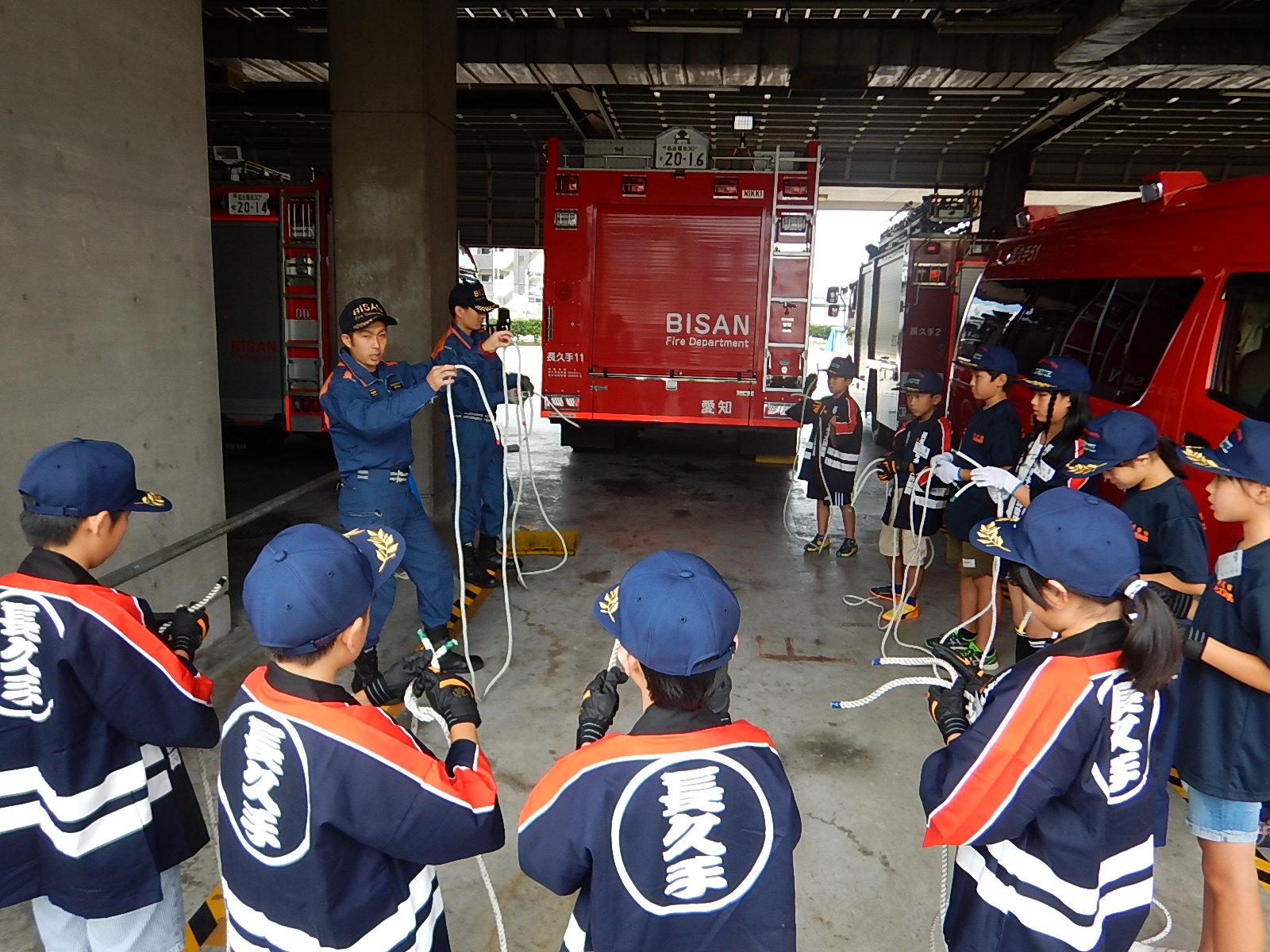 消防車両が駐車してある車庫で消防署員よりロープの結び方を教わるキッズ消防団員たちの写真