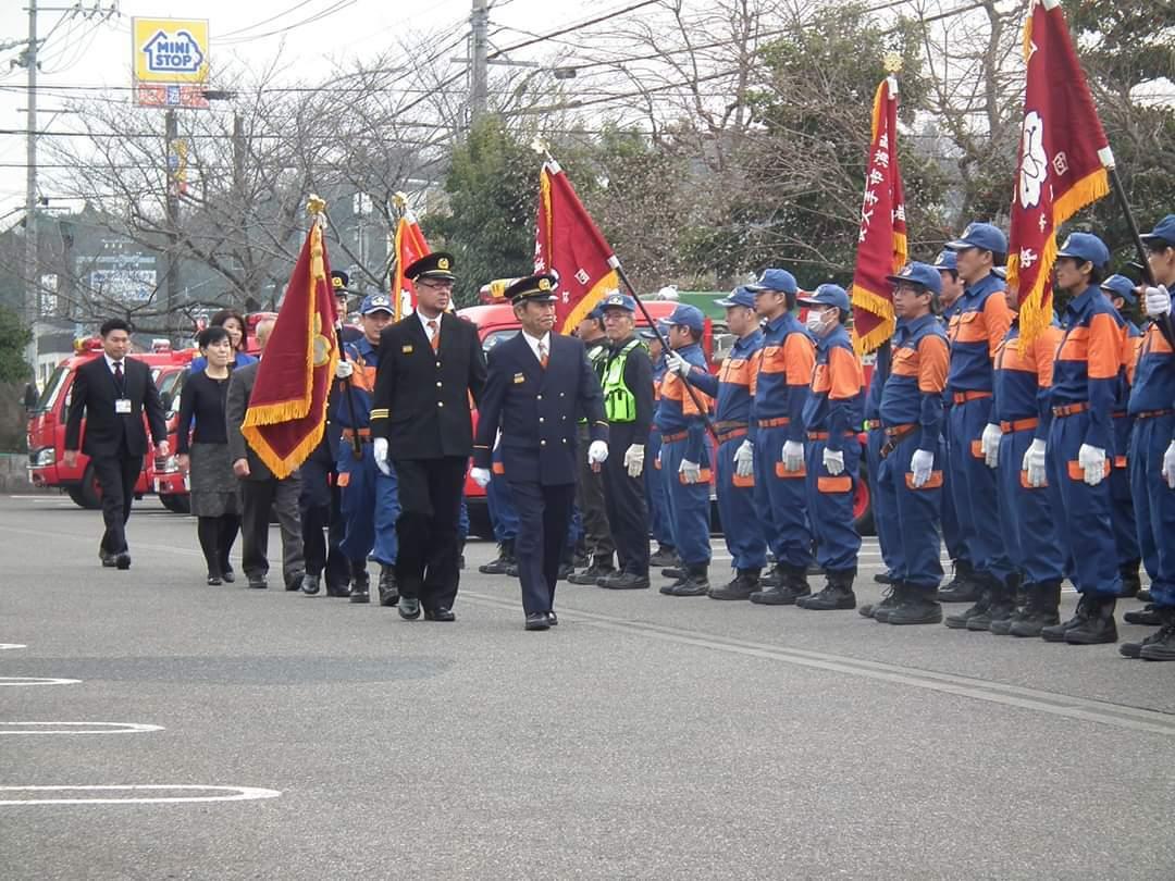 駐車場に旗を持ち整列している消防団員たちの前を団長や来賓たちが歩いている写真