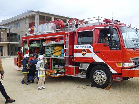 学校の校庭に消防車を展示しているそばで、小さな男の子たちが消防士の服装に着替えている写真
