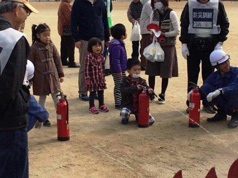 避難訓練に親子で参加した小さな子供たちが、消防団の指導のもと消火器を使っての消火訓練を行っている写真