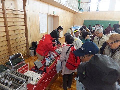 （長小）NTTドコモによる災害用伝言板体験訓練ブースの受付に携帯電話やスマートフォンが並べられており、赤いジャンバーを着たスタッフより説明を受ける参加者たちの写真