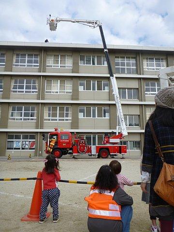 4階建ての校舎の屋上の上まではしごが伸びている消防車による訓練を、小さな女の子と女性が見学している写真
