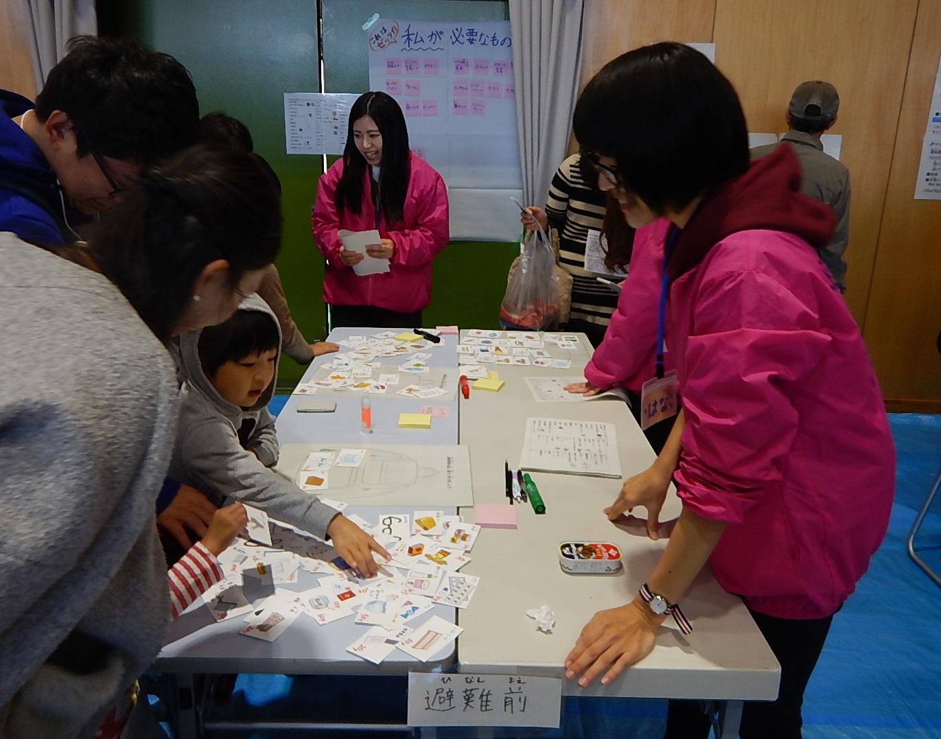 長机の上に広げられたカードを指さしている参加者の子供とその様子を見守っている保護者とピンク色の上着を着た看護学生の写真