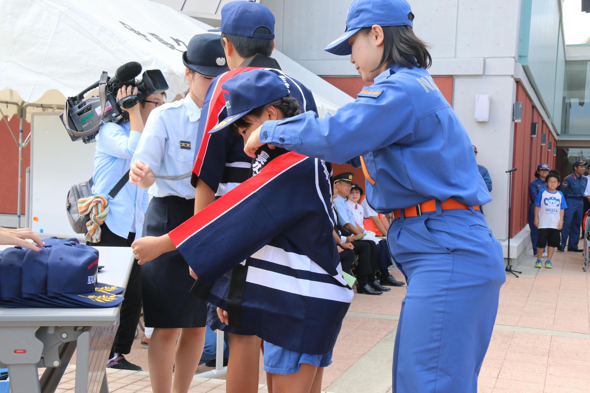 青い制服を着た女性から帽子と法被を着させてもらっている女の子の写真